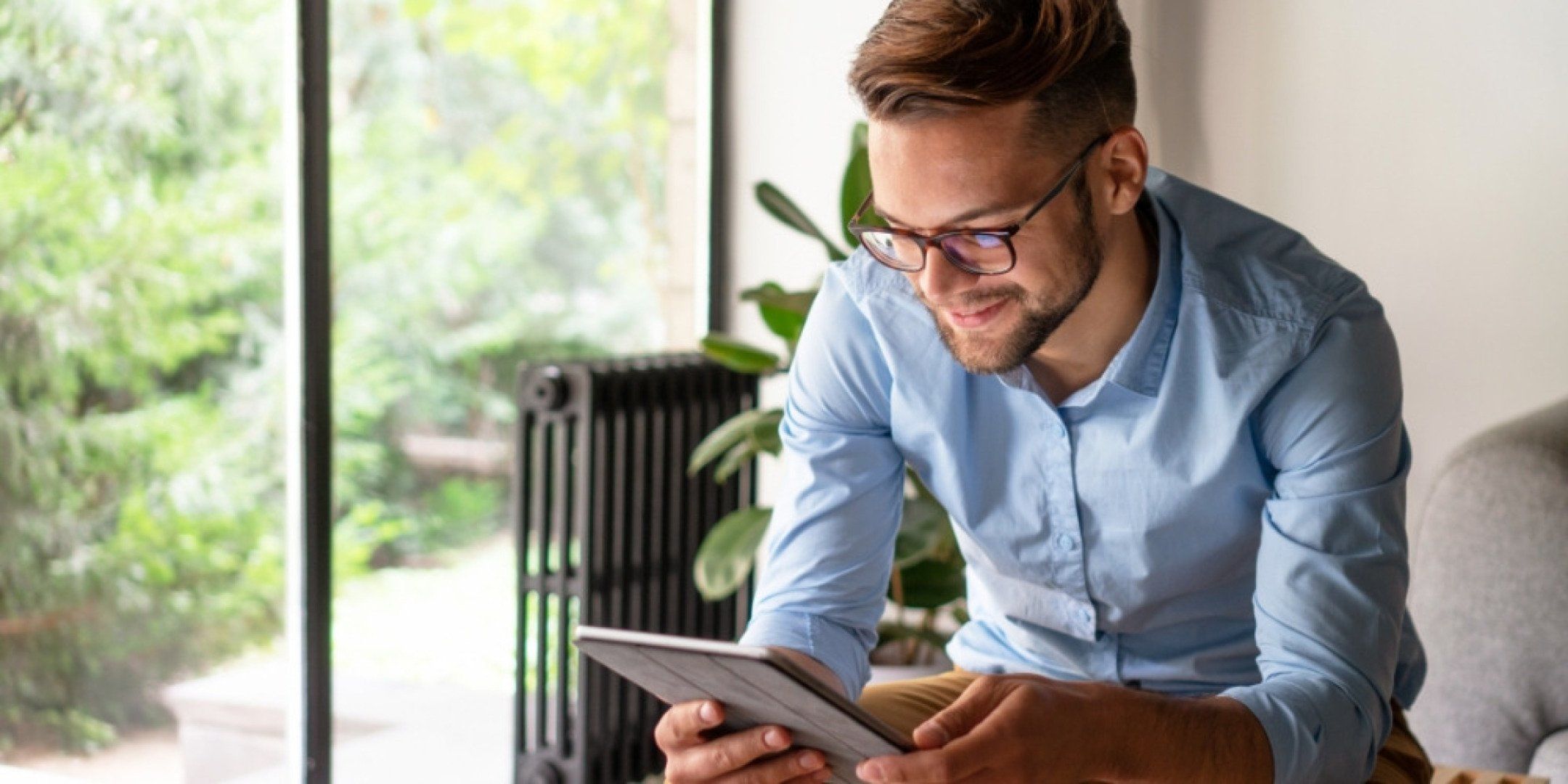 Auf dem Bild ist ein junger Mann zu sehen. Er sitzt auf dem Sofa und schaut auf sein Tablet.
