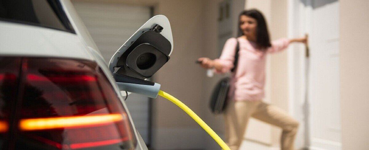Elektroauto mit Ladekabel mit einer Frau im Hintergrund, die auf die Fernbedienung des Fahrzeugs drückt und zur Haustür geht.