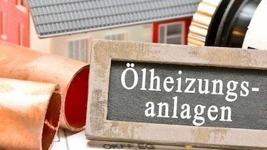 Hier ist ein kleines Haus zu sehen, davor liegen Heizungsrohre, ein Thermostatkopf und eine Tafel mit der Aufschrift „Ölheizungsanlage“.